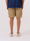 The Sand Recycled Polytech Parachute Short is worn by a model against a plain white background. The image shows a rear three-quarter view of the model from the waist down, highlighting the elasticated waistband with a drawcord and two large rear patch pockets with button flaps. The fabric has a smooth, technical appearance in a light sand color. No mannequin is present in the image.