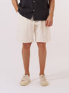 A model wears the Ecru Slub Drill Pleated Track Short, standing against a plain white background. The image shows a front view of the shorts, highlighting the elasticated waistband, the distinct single pleat on each leg, and the textured ecru cotton slub drill fabric. The model is wearing a light-colored shirt tucked into the shorts.