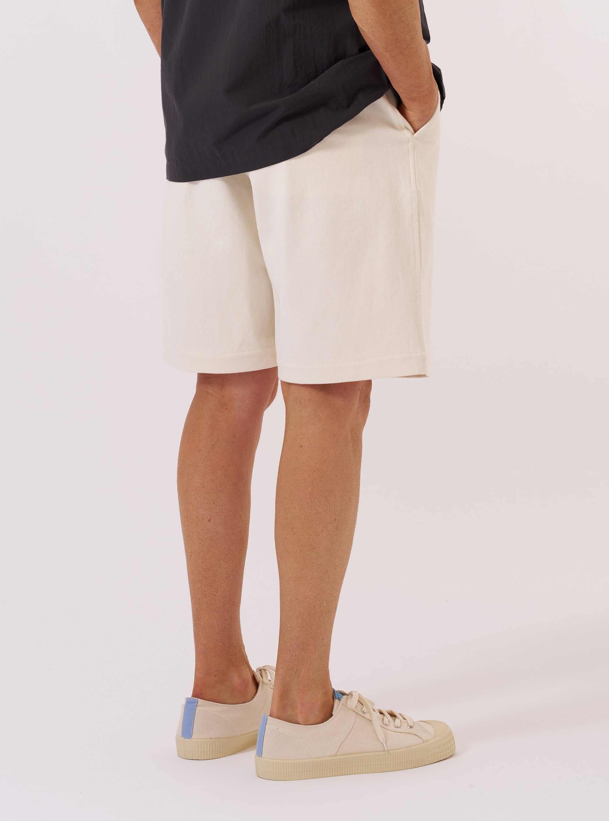A model wears the Ecru Slub Drill Pleated Track Short, shown from the waist down from a rear three-quarter angle against a plain white background. The shorts feature a relaxed fit, visible front pleats, and a single patch pocket on the right rear side. The textured ecru cotton slub drill fabric and straight hem are prominent features.