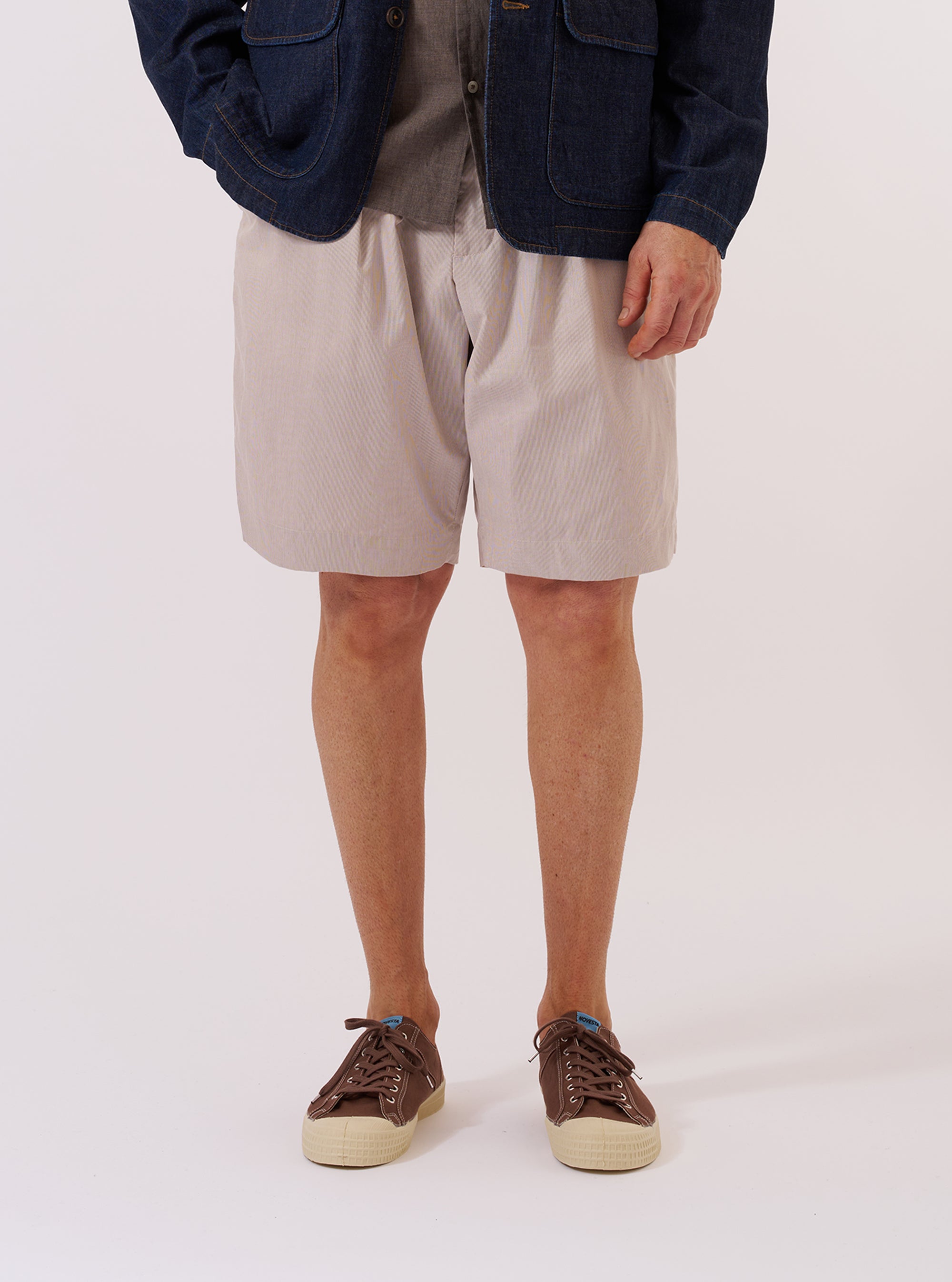 A model wears the Brown Kennedy Stripe Pleated Track Short against a plain white background. The shorts feature a vertical brown and cream striped pattern, a pleated front, and a relaxed fit. The model is shown from the waist down, highlighting the drape of the fabric and the mid-thigh length of the garment.