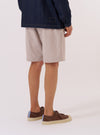 A model wears the Brown Kennedy Stripe Pleated Track Short, shown from the waist down against a plain white background. The shorts feature a vertical brown and white stripe pattern, a pleated front, and a relaxed fit. The model is standing with their hands in their pockets.