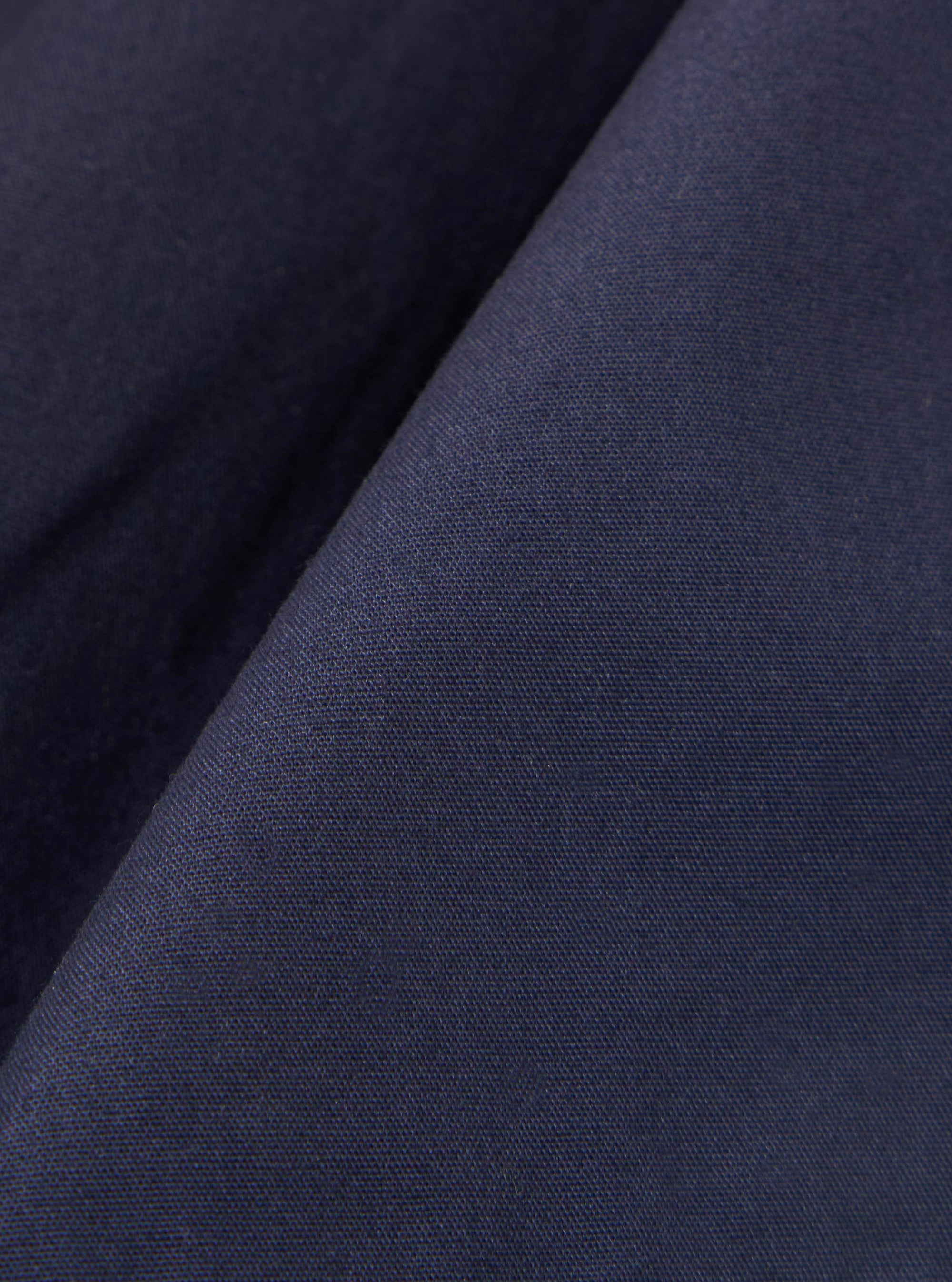 A close-up fabric swatch of the Navy Peached Poplin Pleated Track Short highlights the smooth, matte texture and deep navy blue color of the peached poplin material. The fabric fills the frame, showcasing its fine weave and soft finish.