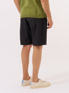 A model is wearing the Black Recycled Polytech Pleated Track Short, shown from the waist down against a plain white background. The shorts feature a relaxed fit, prominent front pleats, and an elasticated waistband with integrated belt loops. The garment is constructed from a smooth, black recycled polytech fabric with a clean, utilitarian appearance.