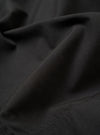 A close-up fabric swatch of the Black Recycled Polytech Pleated Track Short is displayed, highlighting the solid black color and the smooth, technical texture of the recycled polytech material.