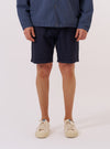A model wears the Navy Dot Waffle Simple Short while standing against a plain white background. The image shows a front view of the shorts, highlighting the elasticated waistband and the textured dot waffle cotton fabric. The model is wearing the shorts with a white T-shirt tucked in and white socks.