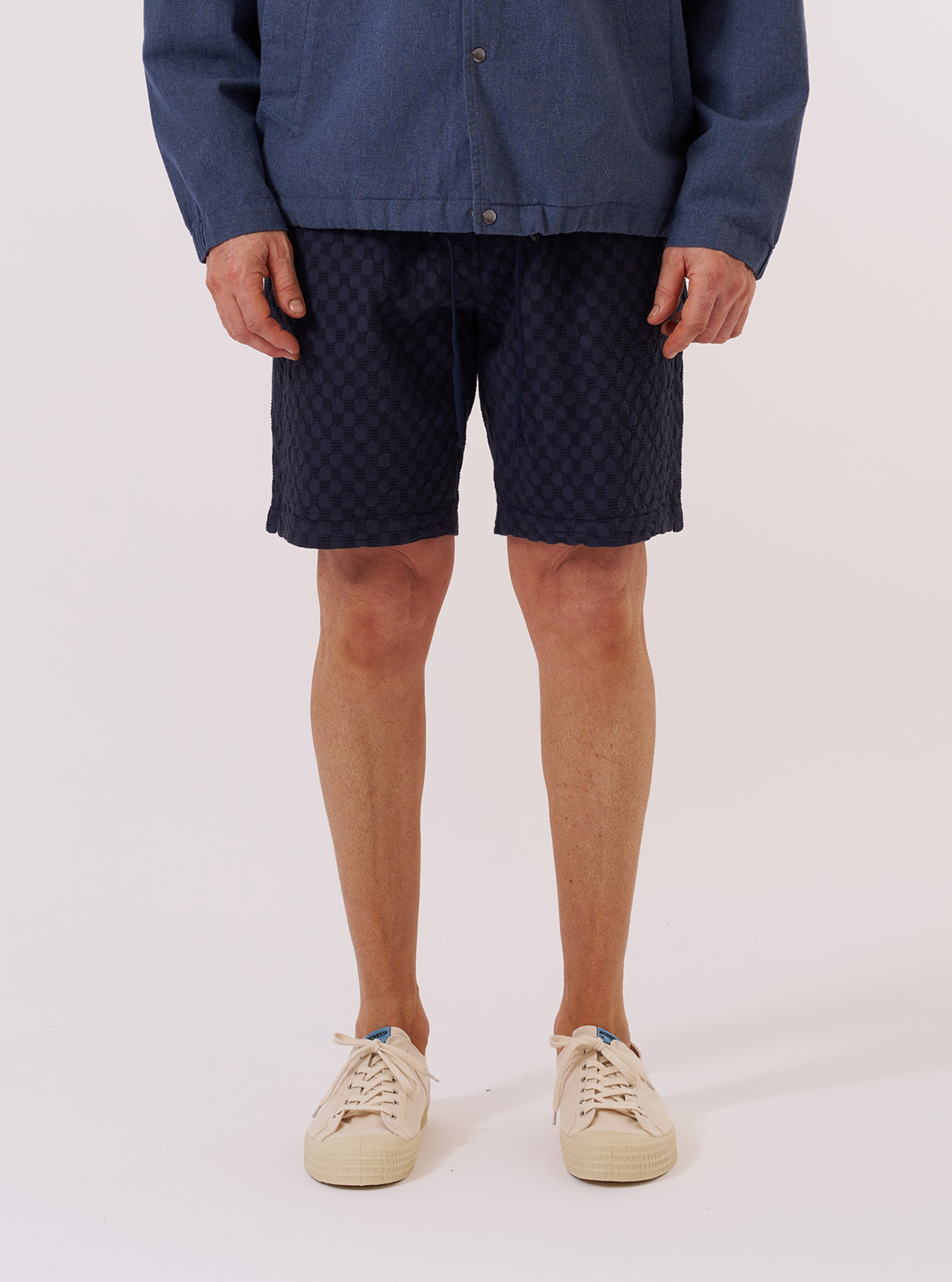A model wears the Navy Dot Waffle Simple Short while standing against a plain white background. The image shows a front view of the shorts, highlighting the elasticated waistband and the textured dot waffle cotton fabric. The model is wearing the shorts with a white T-shirt tucked in and white socks.