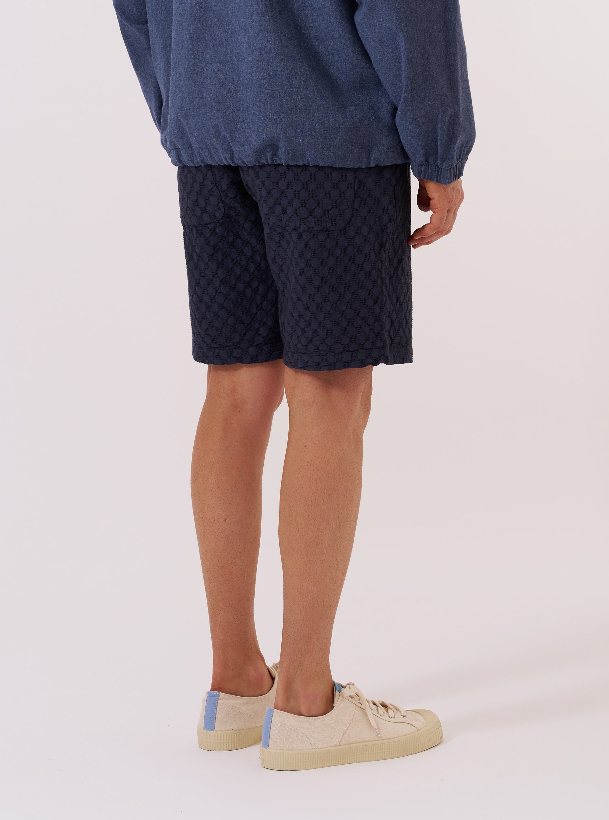 The Navy Dot Waffle Simple Short is worn by a model against a plain white background. The image shows a rear view of the shorts, highlighting the elasticated waistband and the textured dot waffle fabric in navy blue. The model is also wearing a white T-shirt and white socks.