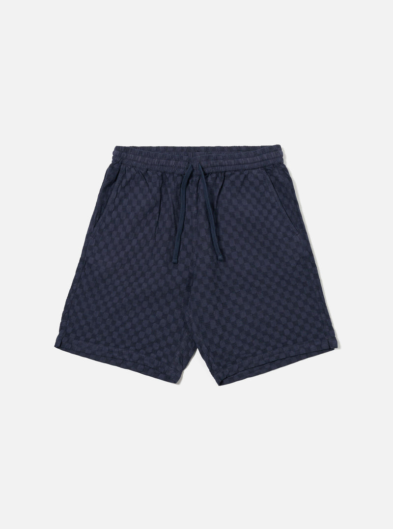 The Navy Dot Waffle Simple Short is shown as a flat lay against a plain white background. The shorts feature a textured waffle knit fabric in navy blue with a subtle dot pattern, an elasticated waistband with a matching drawstring, and two side pockets. The design is simple with a relaxed fit and straight hems.