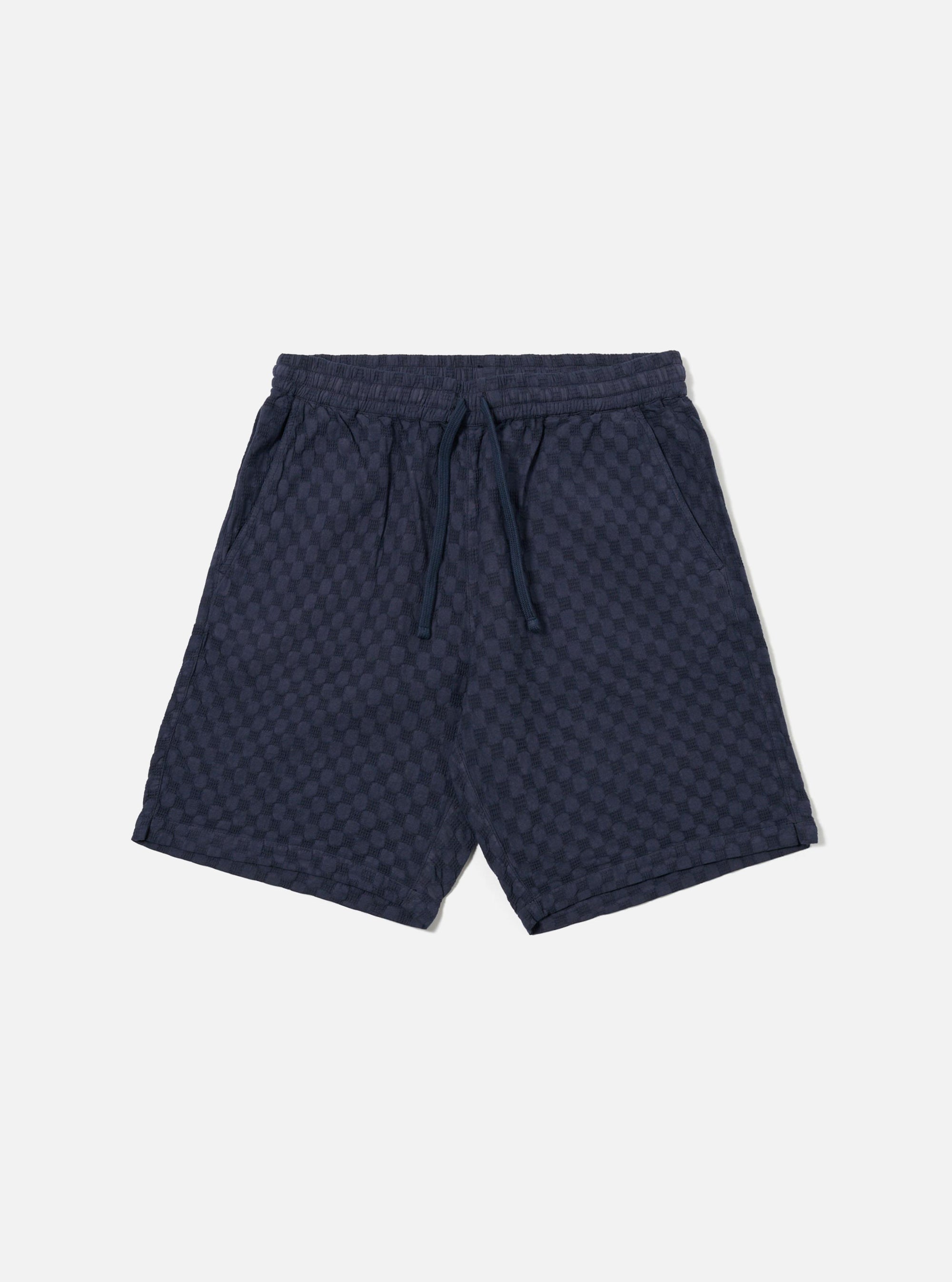 The Navy Dot Waffle Simple Short is shown as a flat lay against a plain white background. The shorts feature a textured waffle knit fabric in navy blue with a subtle dot pattern, an elasticated waistband with a matching drawstring, and two side pockets. The design is simple with a relaxed fit and straight hems.