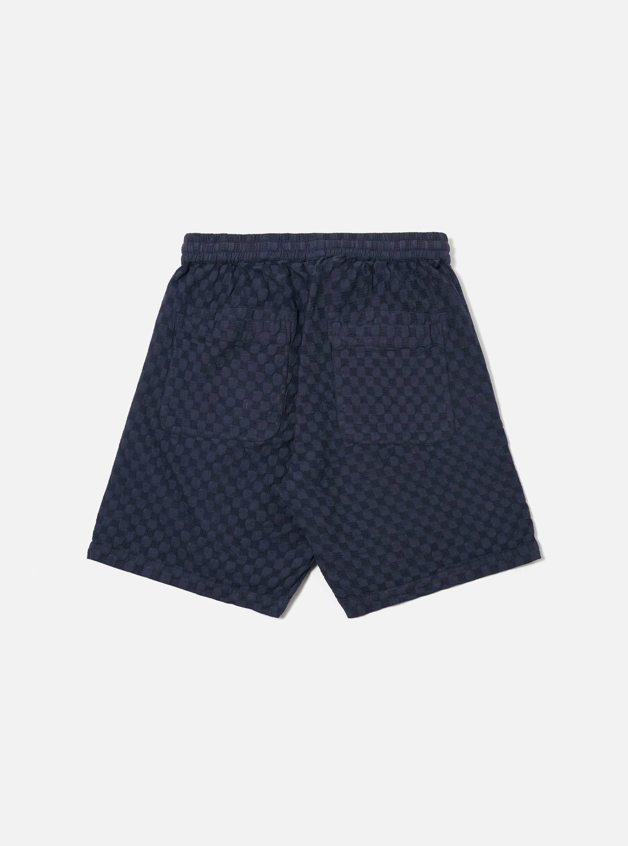 The Navy Dot Waffle Simple Short is shown from the back as a flat lay against a plain white background. The image highlights the elasticated waistband and a single rear patch pocket on the right side. The navy blue fabric features a distinct, textured waffle knit pattern with small dot-like indentations.
