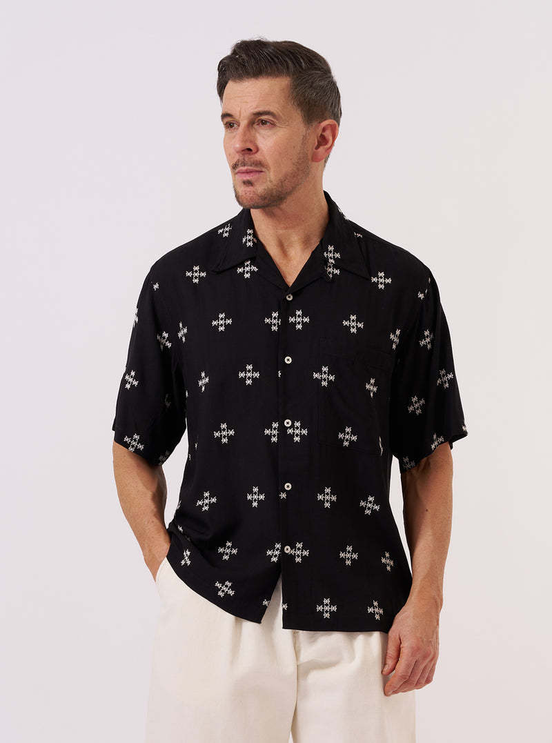 A model wears the Black Embroidered Diamond Camp Shirt over a white top, standing against a plain, neutral background. The black viscose shirt features a relaxed camp collar, short sleeves, and an intricate white diamond embroidery pattern throughout. One large chest pocket is visible on the front.