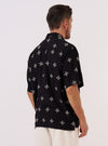 A model is wearing the Black Embroidered Diamond Camp Shirt, shown from the back against a plain white background. The shirt features a relaxed fit with short sleeves and a straight hem that is slightly longer in the back with side splits. A subtle embroidered diamond pattern is visible across the black fabric, particularly around the back yoke area. No mannequin is present in the image.