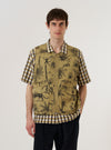 The Sand Jungle Toile Twill 4 Pocket Gilet is worn by a model and shown against a plain white background. The gilet is made from a sand-colored twill fabric featuring an intricate dark jungle toile print throughout. It is designed with four large patch pockets on the front and a central button-down closure. The model is wearing the gilet layered over a neutral-colored long-sleeve top.
