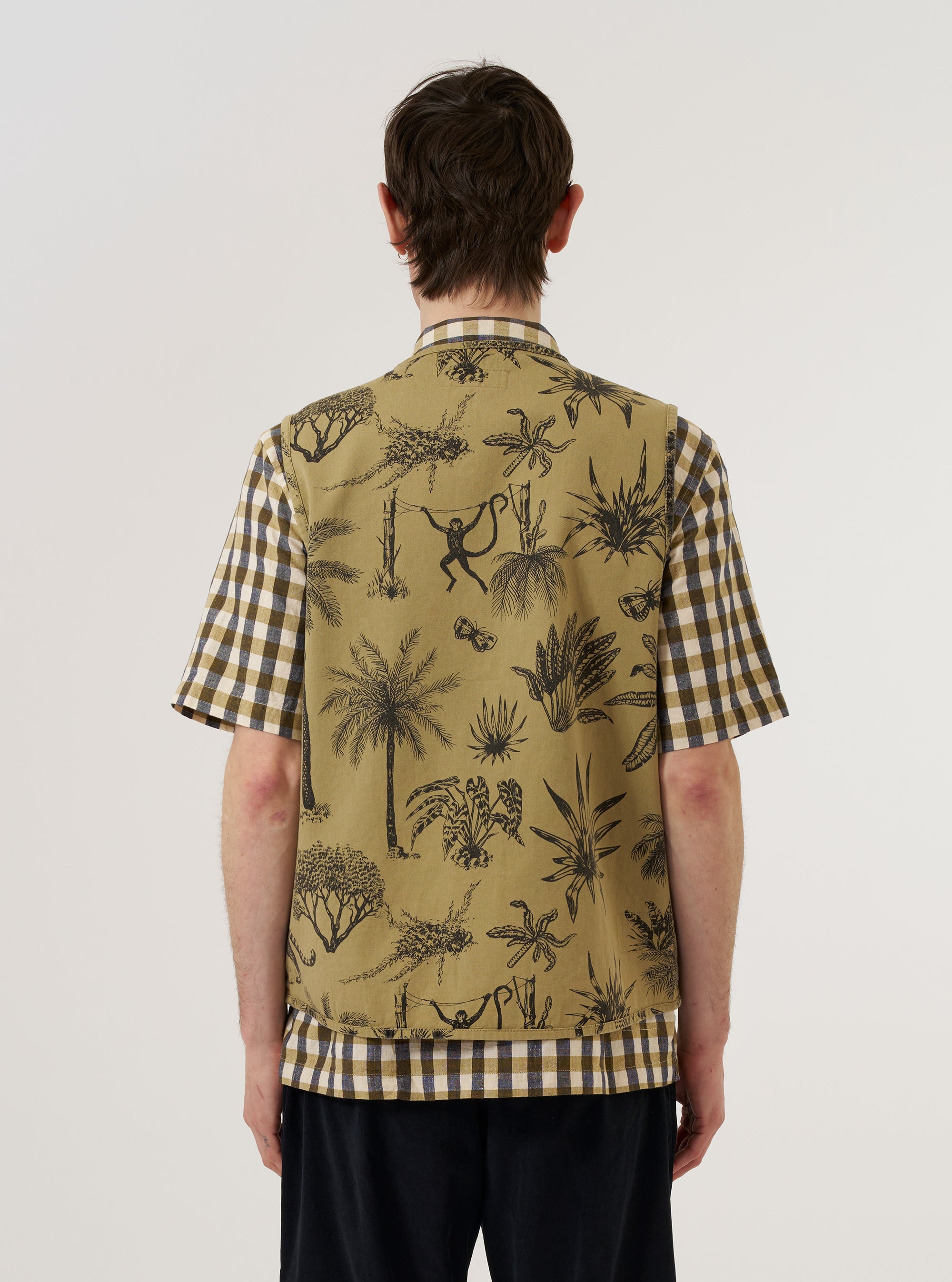 A model wears the Sand Jungle Toile Twill 4 Pocket Gilet, shown from the back against a plain white background. The garment features an all-over jungle toile print with dark botanical illustrations on a sand-colored base. The sleeveless design and straight-cut back are clearly visible as the model stands facing away from the camera.