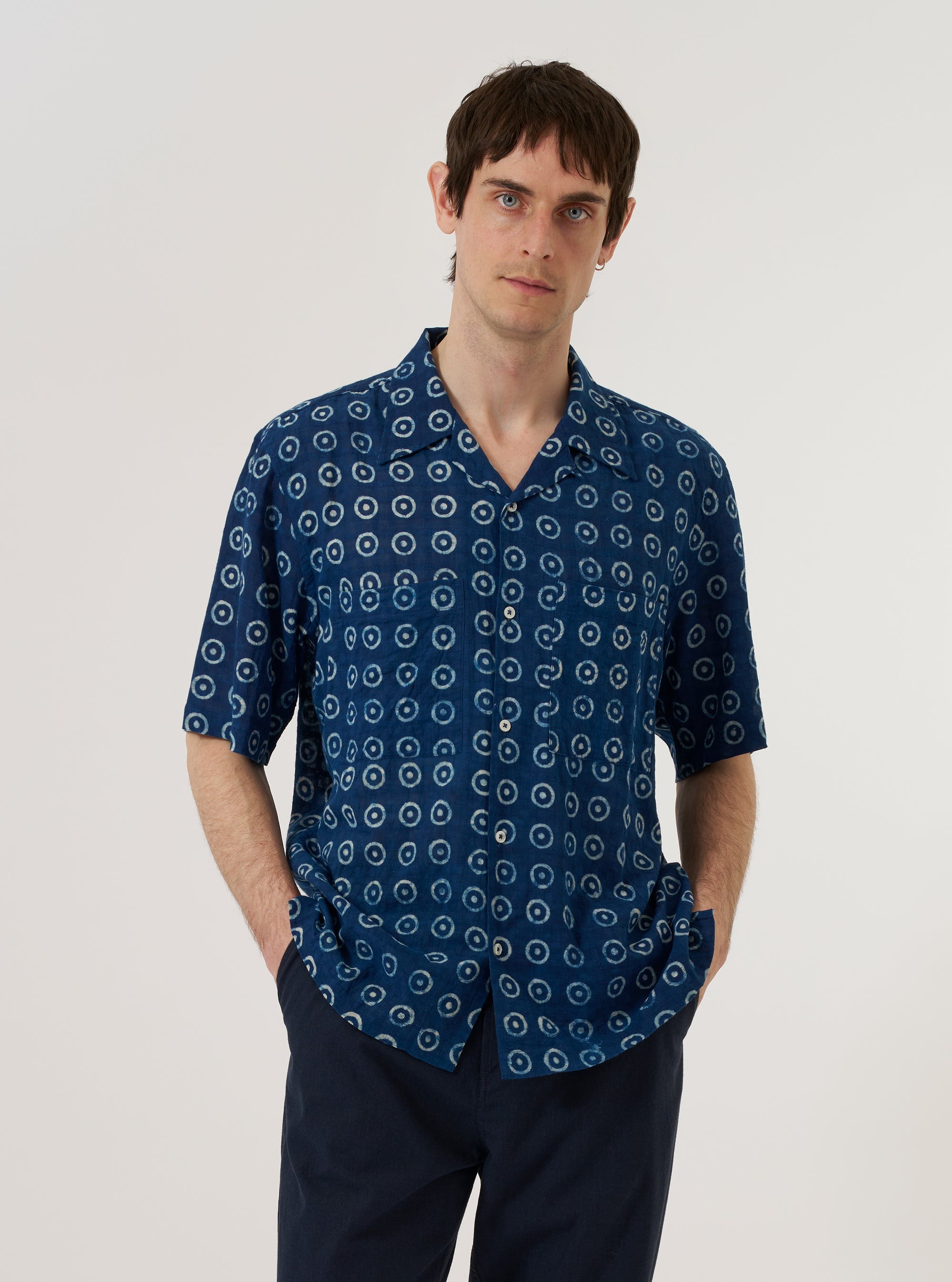 A model is wearing the Indigo Block Print Circles Camper Shirt over a white t-shirt, standing against a plain white background. The shirt features a camp collar, a single chest pocket, and a central button-down front. The indigo blue fabric is decorated with a repeating white hand-block printed circle pattern. The model is shown from the chest up in a medium shot.