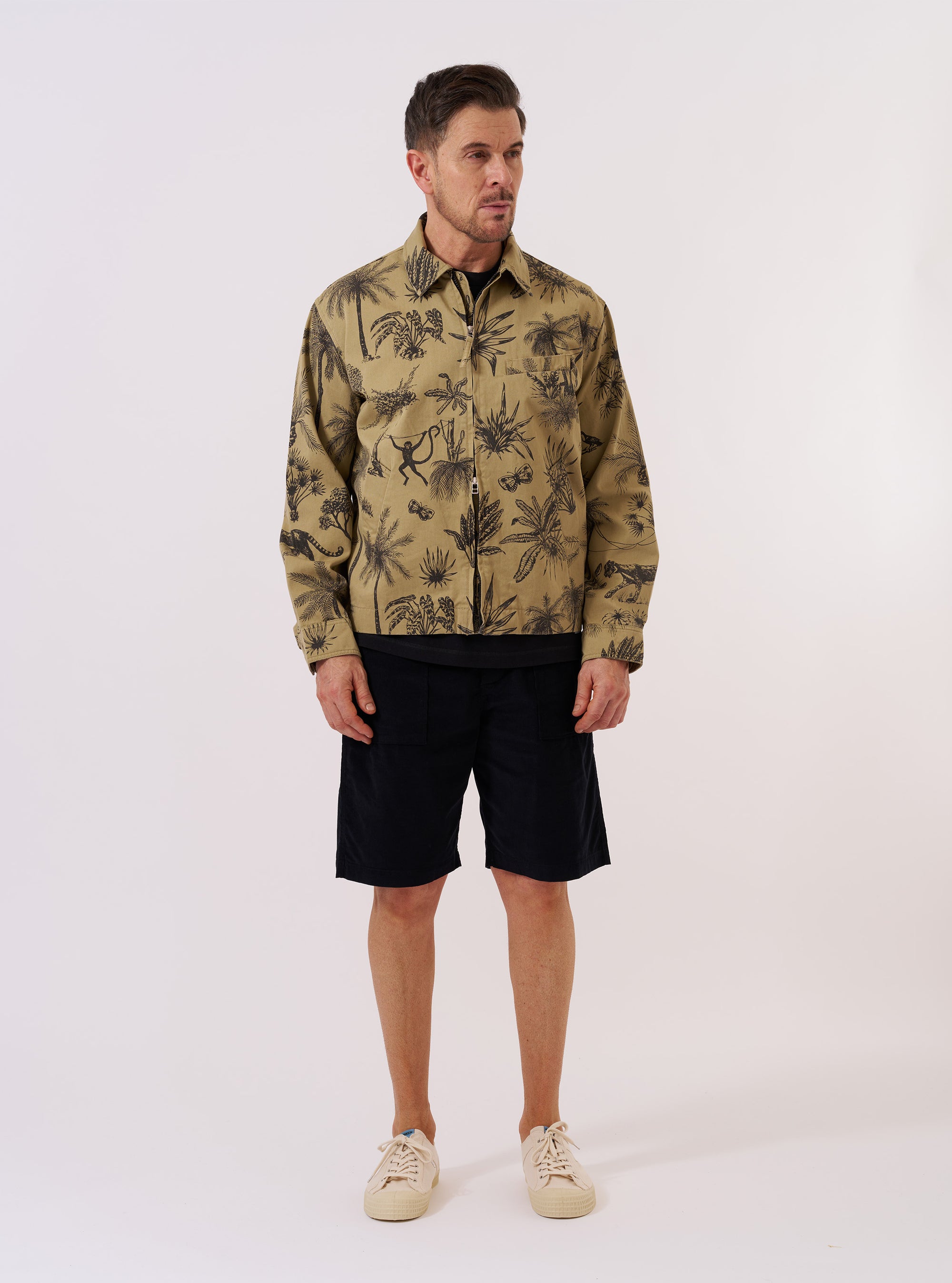 The Sand Jungle Toile Twill Windcheater Jacket is presented as a flat lay against a plain white background. The jacket features an all-over dark jungle toile print on a sand-colored base, a classic collar, and a central zip-front closure. Two large slanted patch pockets are prominent on the front.