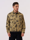 A model wears the Sand Jungle Toile Twill Windcheater Jacket against a plain white background. The jacket features an all-over dark jungle toile print on a sand-colored base, a classic collar, and a full-length silver zipper. The model is shown from the chest up with the jacket zipped, highlighting the intricate pattern of tropical foliage and animals.