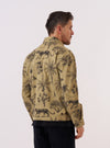 A model wears the Sand Jungle Toile Twill Windcheater Jacket, shown from the rear three-quarter angle against a plain white background. The jacket features an all-over dark jungle toile print on a sand-colored base, a simple collar, and a straight hem. The model is wearing the jacket over a light-colored top.