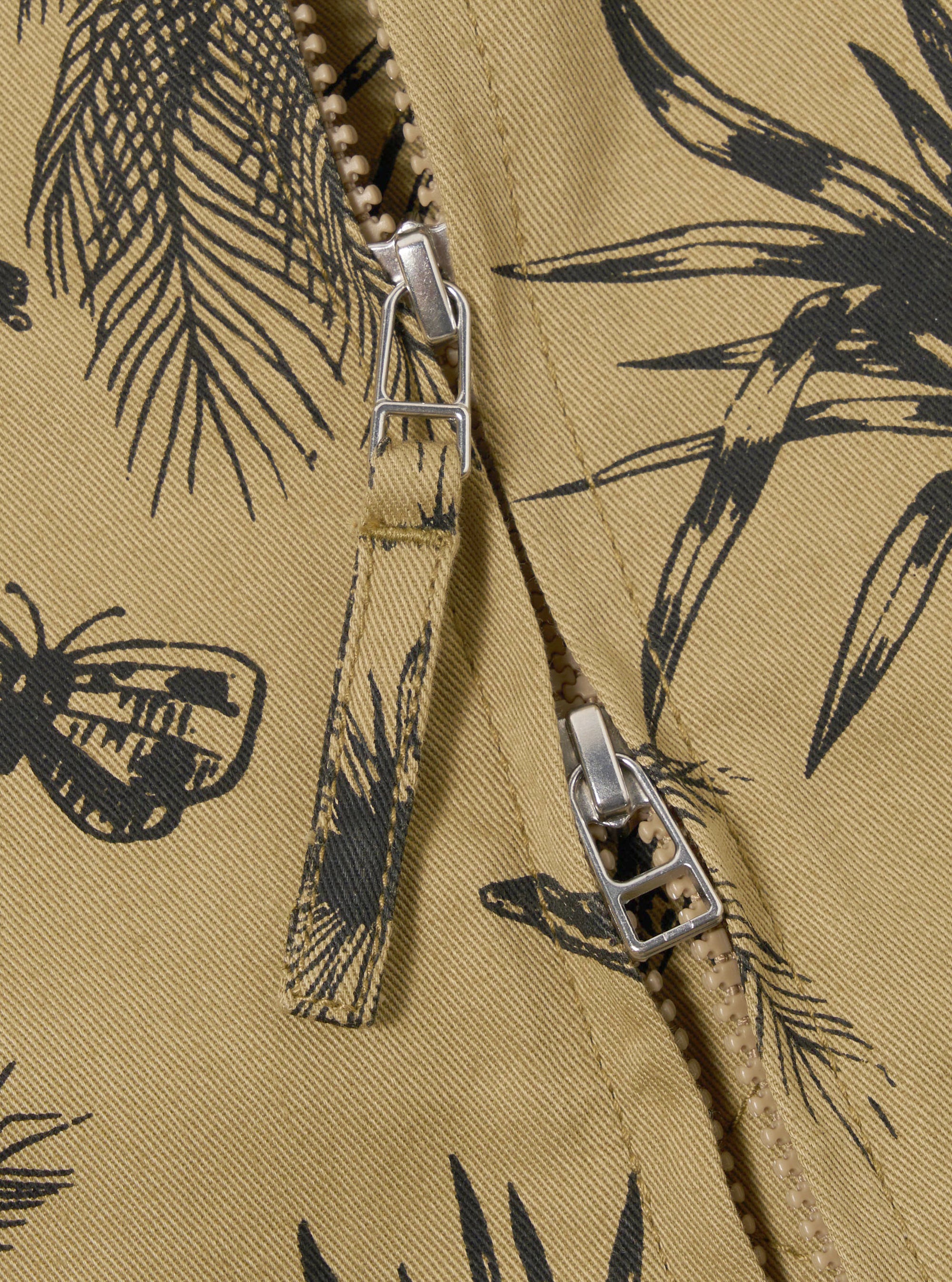 The Sand Jungle Toile Twill Windcheater Jacket is shown in a detail view as a flat lay against a plain white background. The image focuses on the lower half of the jacket, highlighting the intricate dark jungle toile print on the sand-colored twill fabric and the clean finish of the straight hem.