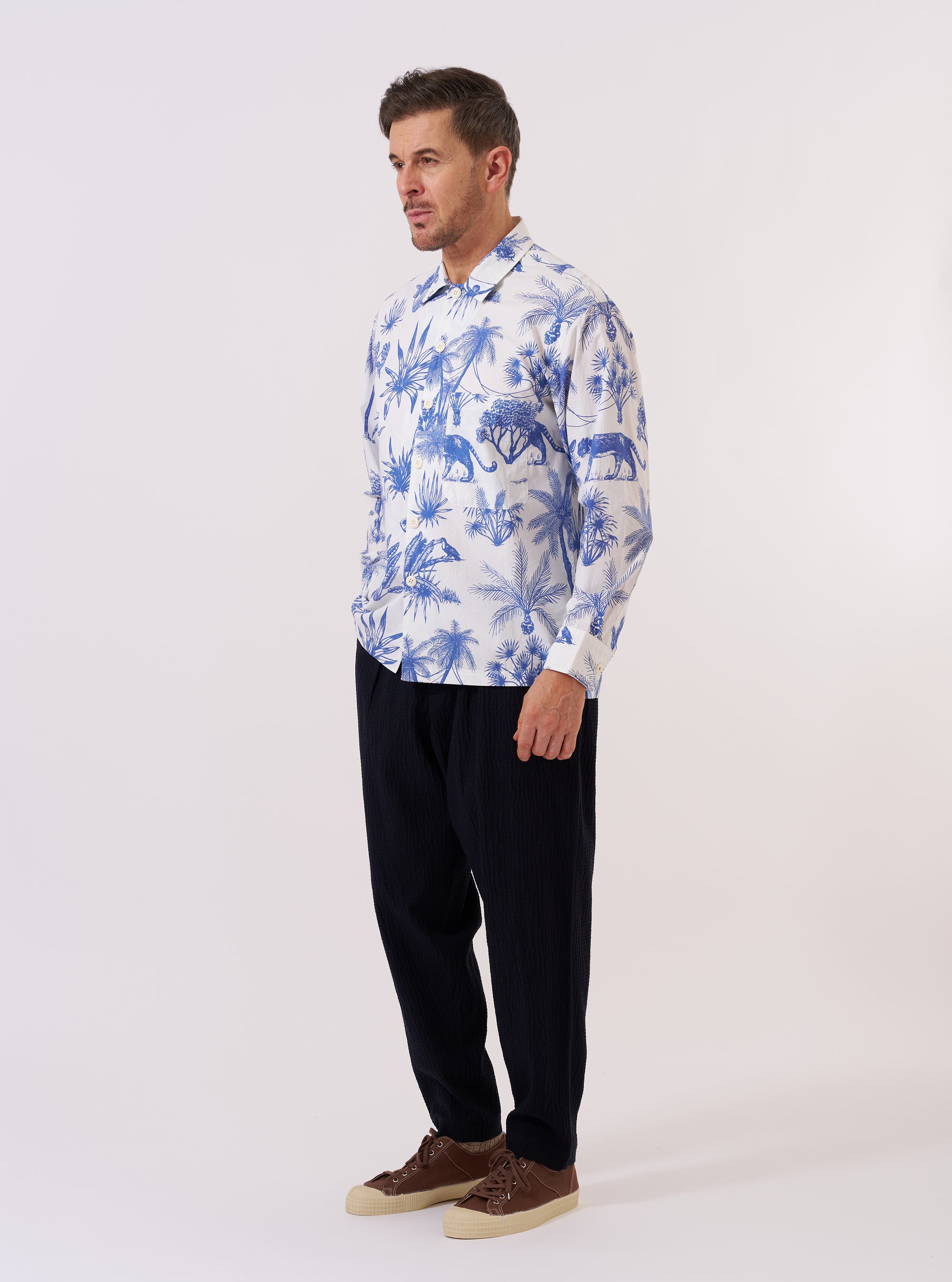 The White Jungle Toile Poplin Fatigue Shirt is shown as a flat lay against a plain white background. The shirt features an all-over jungle toile print with dark botanical and animal illustrations on a white base. Key details include a one-piece collar, a single large square chest patch pocket on the left side, and a central button-down front with oversized white buttons. The shirt has a straight hem and long sleeves with reinforced cuffs.
