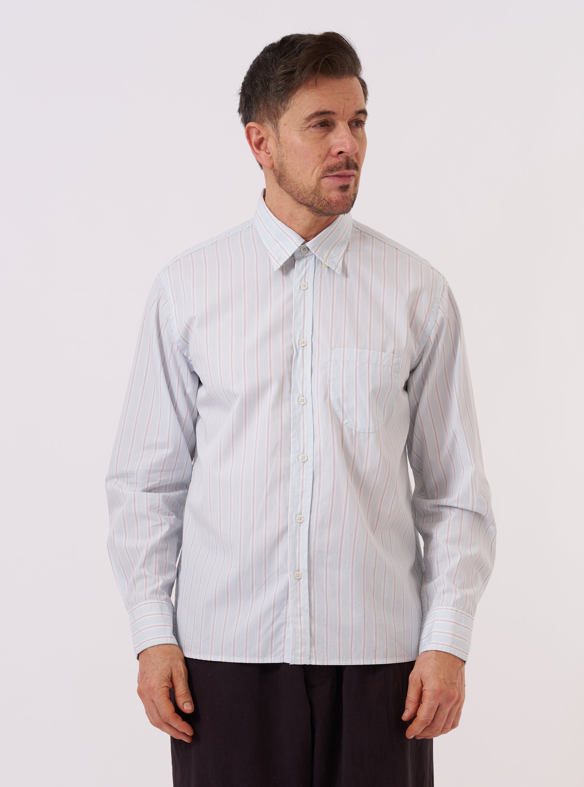 The Sky Urban Stripe Lazy Day Shirt is worn by a model standing against a plain white background. The shirt features a light blue and white vertical stripe pattern, a button-down collar, and a single asymmetrical chest pocket. The model is wearing the shirt buttoned up, showcasing its relaxed fit.