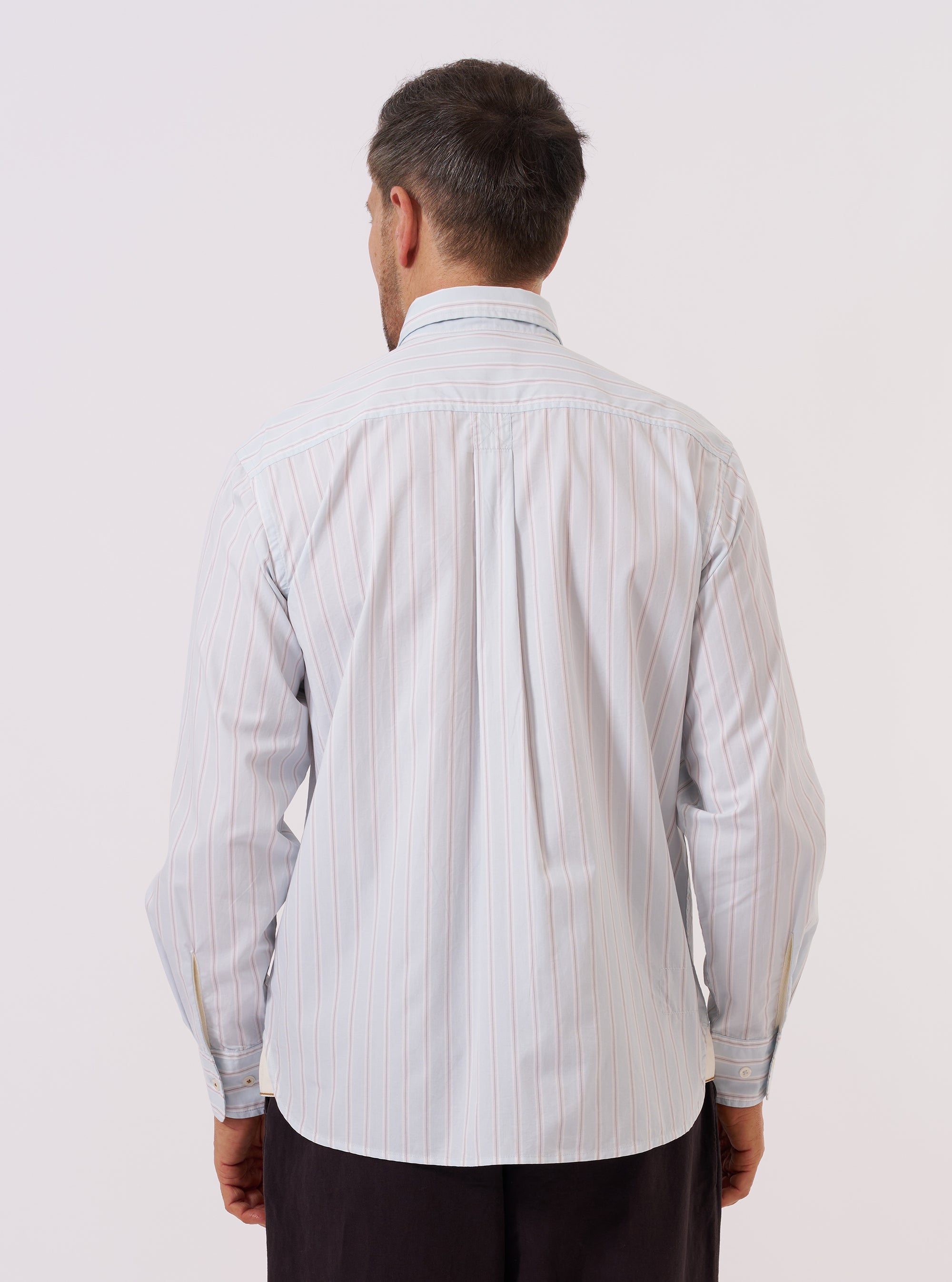 A model wears the Sky Urban Stripe Lazy Day Shirt against a plain white background. The image shows the shirt from the back, highlighting the vertical blue and white stripes and the straight hem. The model is shown from the head down to the waist, wearing the shirt tucked into dark trousers.