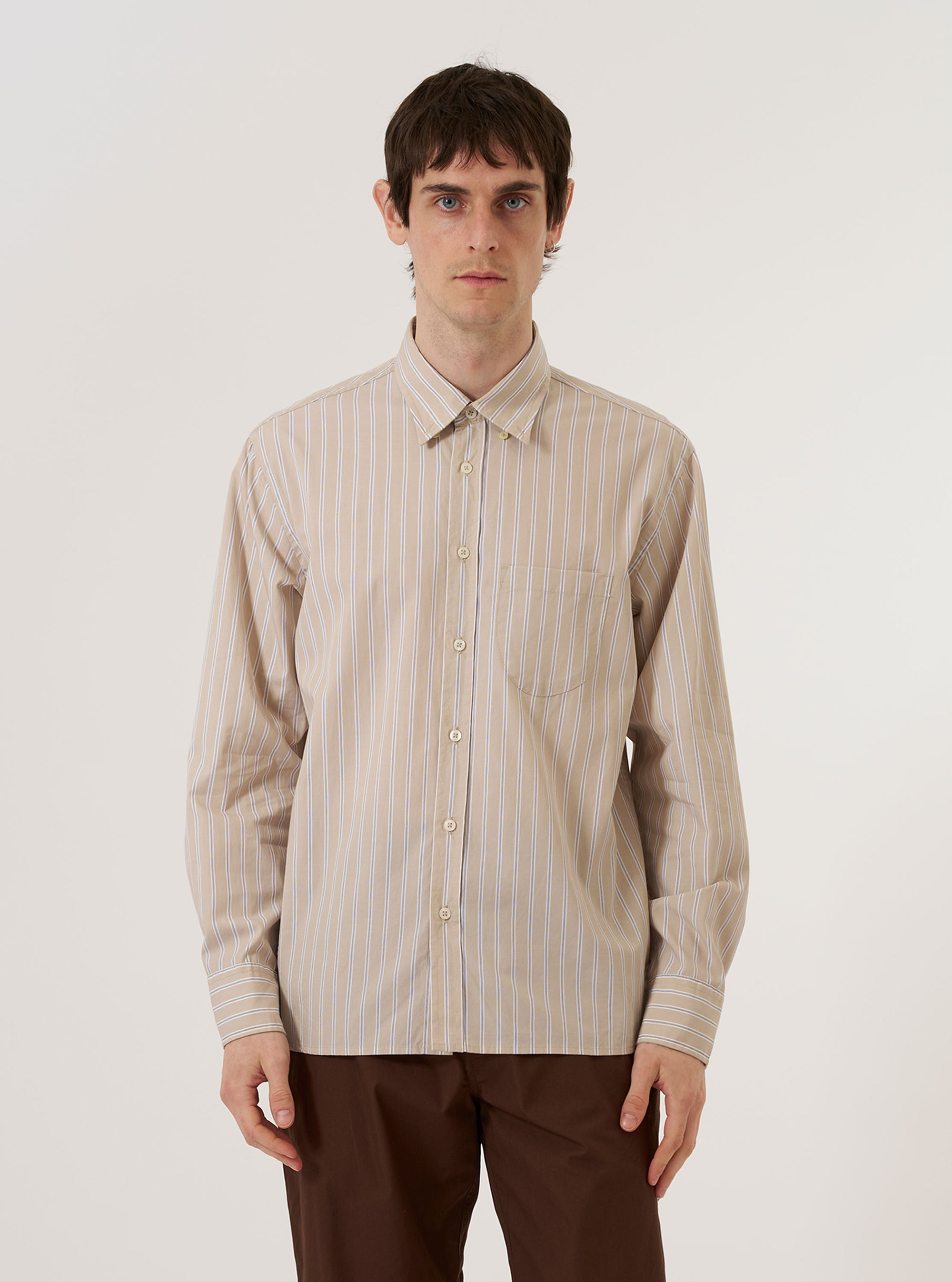 A model wears the Sand Urban Stripe Lazy Day Shirt while standing against a plain white background. The shirt features a vertical striped pattern in sand and white, a button-down front, and a single chest pocket. The model is wearing the shirt untucked over a light-colored undershirt.
