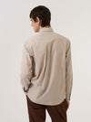 The Sand Urban Stripe Lazy Day Shirt is worn by a model against a plain white background. The image shows the model from the back, highlighting the shirt's relaxed fit, back yoke, and box pleat. The fabric features a fine vertical stripe pattern in sand and stone colors. The model is also wearing dark trousers.