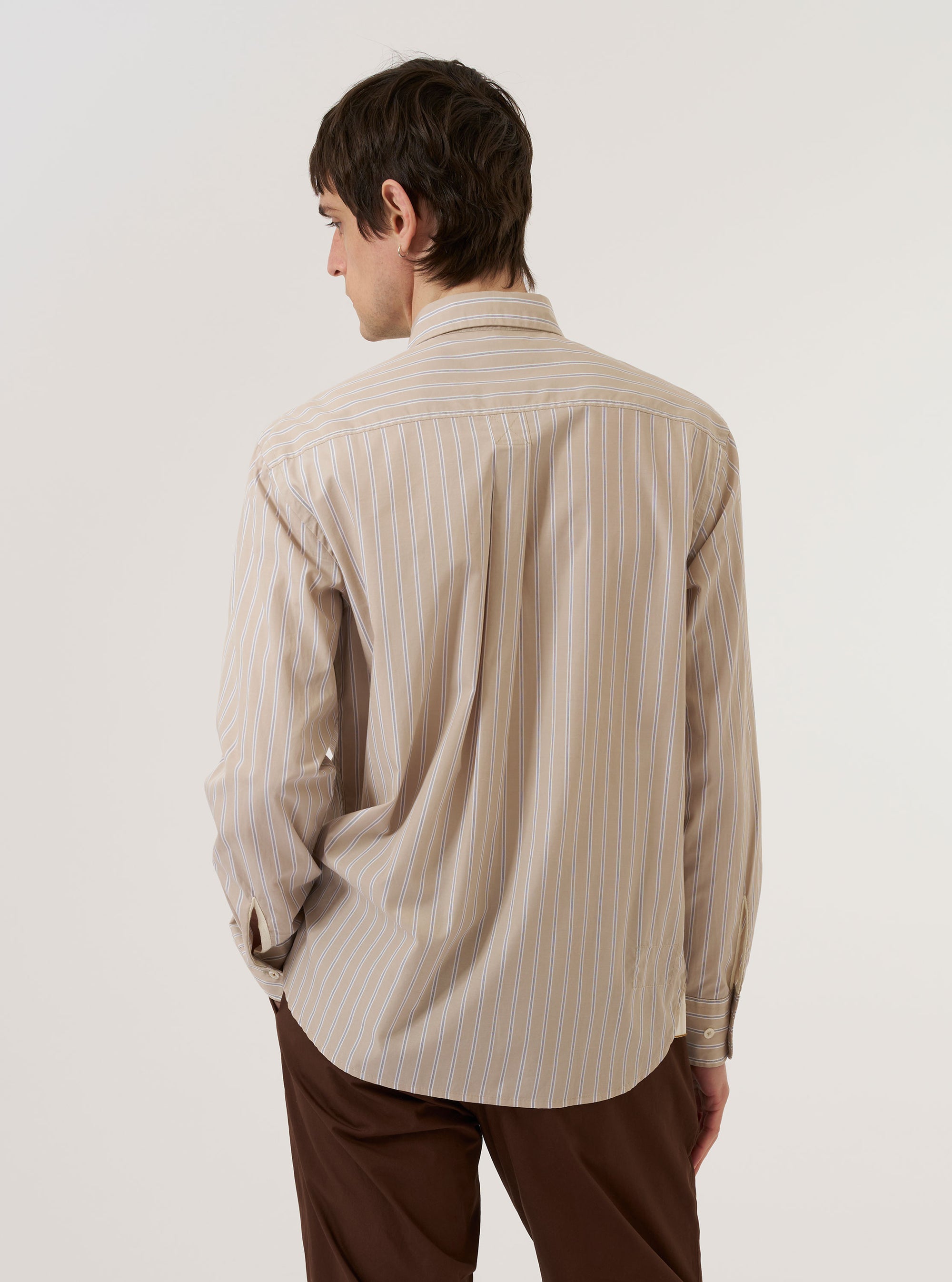 The Sand Urban Stripe Lazy Day Shirt is worn by a model against a plain white background. The image shows the model from the back, highlighting the shirt's relaxed fit, back yoke, and box pleat. The fabric features a fine vertical stripe pattern in sand and stone colors. The model is also wearing dark trousers.