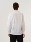 A model wears the White Poplin Dwight Overshirt against a plain white background, shown from a rear three-quarter angle. The image highlights the garment's clean, white poplin fabric, a straight hem, and a simple, structured back. The overshirt features a relaxed fit and a camp collar. No mannequin is present in the image.