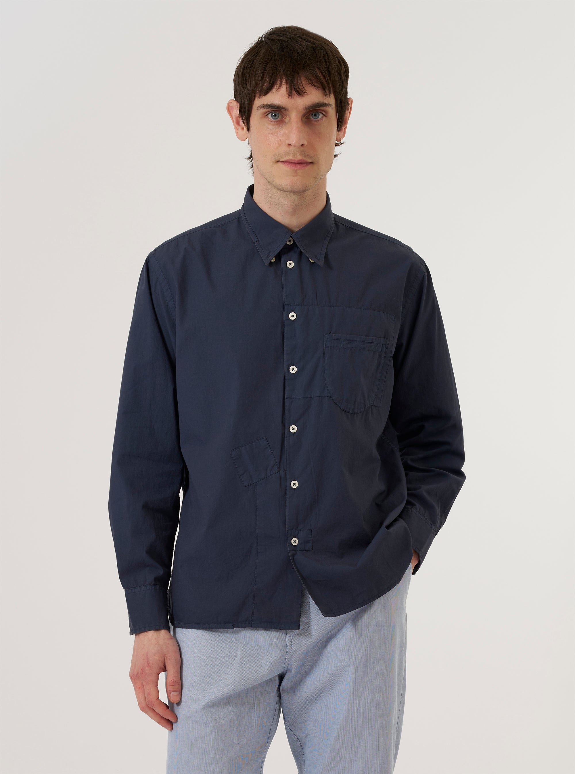 A model wears the Navy Poplin Patched Lazy Day Shirt while standing against a plain white background. The shirt features a button-down front, a classic collar, and tonal navy patches on the chest and body. The model is shown from the waist up, with the shirt worn over a white T-shirt.
