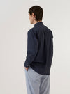 A model wears the Navy Poplin Patched Lazy Day Shirt against a plain white background. The model is shown from the waist up and from a rear three-quarter angle, highlighting the back yoke with its central box pleat and the shirt's relaxed fit. The garment features long sleeves with buttoned cuffs and is crafted from a dark navy cotton poplin fabric with visible patch detailing.