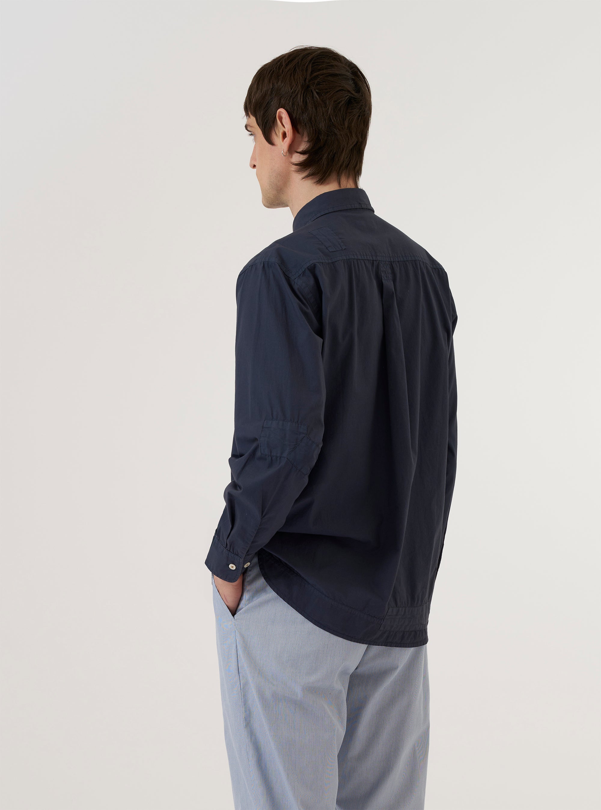 A model wears the Navy Poplin Patched Lazy Day Shirt against a plain white background. The model is shown from the waist up and from a rear three-quarter angle, highlighting the back yoke with its central box pleat and the shirt's relaxed fit. The garment features long sleeves with buttoned cuffs and is crafted from a dark navy cotton poplin fabric with visible patch detailing.