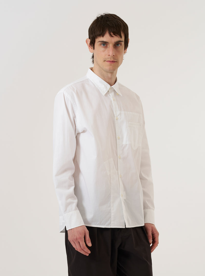 A model wears the White Poplin Patched Lazy Day Shirt while standing against a plain white background. The shirt is shown from the front with the top button unfastened, highlighting its relaxed fit and classic simple collar. The design features a prominent asymmetrical chest pocket and a clean, lightweight cotton poplin fabric. The model's hands are placed in their pockets, and the shirt is worn untucked.