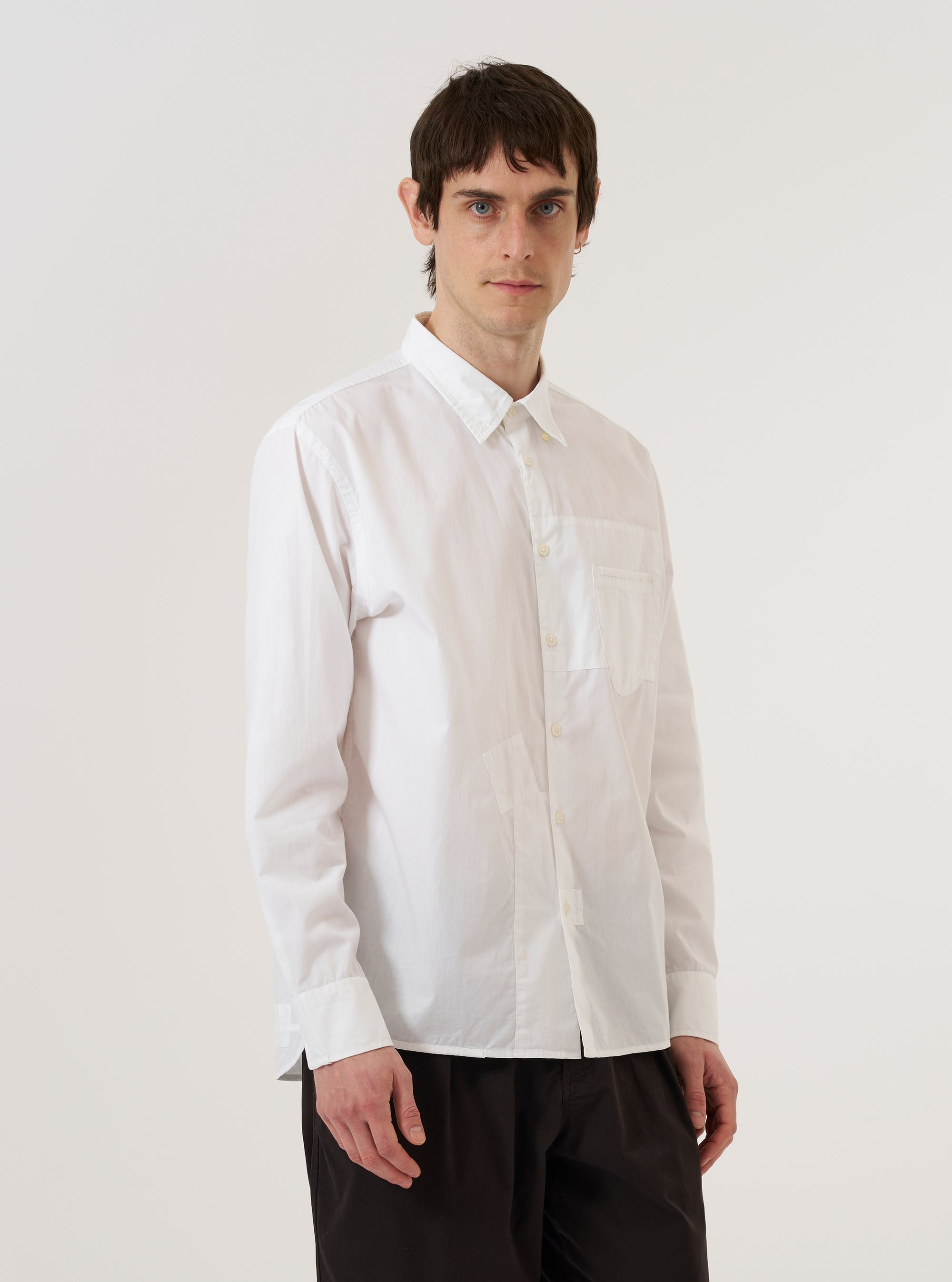 A model wears the White Poplin Patched Lazy Day Shirt while standing against a plain white background. The shirt is shown from the front with the top button unfastened, highlighting its relaxed fit and classic simple collar. The design features a prominent asymmetrical chest pocket and a clean, lightweight cotton poplin fabric. The model's hands are placed in their pockets, and the shirt is worn untucked.