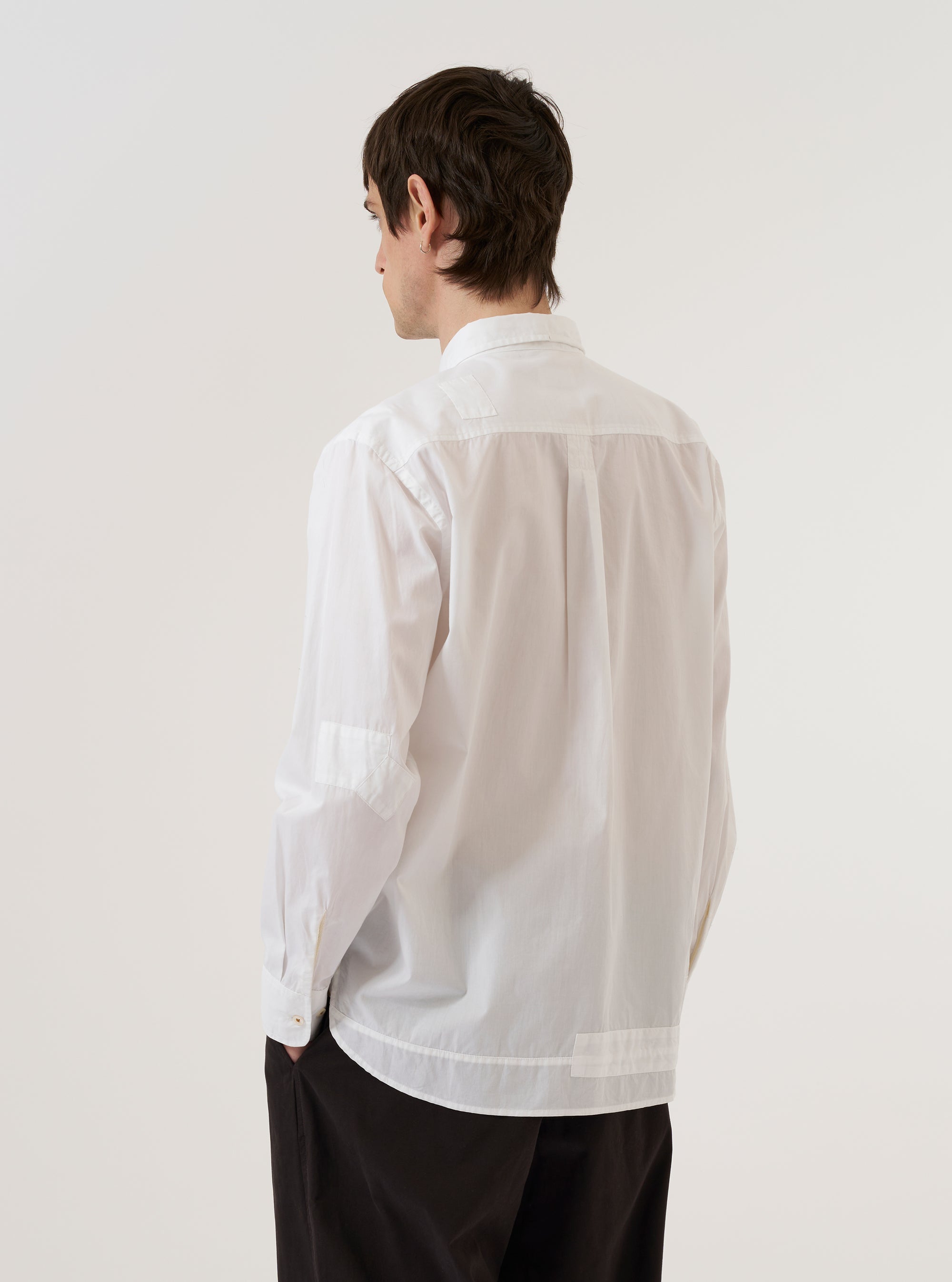 A model wears the White Poplin Patched Lazy Day Shirt against a plain white background. The shirt features a relaxed fit, a button-down front with tonal buttons, and prominent rectangular fabric patches on the chest. The model is shown from the waist up, wearing the crisp white poplin garment tucked into olive green trousers.