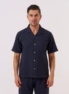A model wears the Navy Dot Waffle Road Shirt against a plain white background. The short-sleeve shirt is shown unbuttoned over a white t-shirt, highlighting its camp collar, single chest patch pocket, and straight hem. The navy blue cotton fabric features a distinct rounded waffle-knit texture throughout.