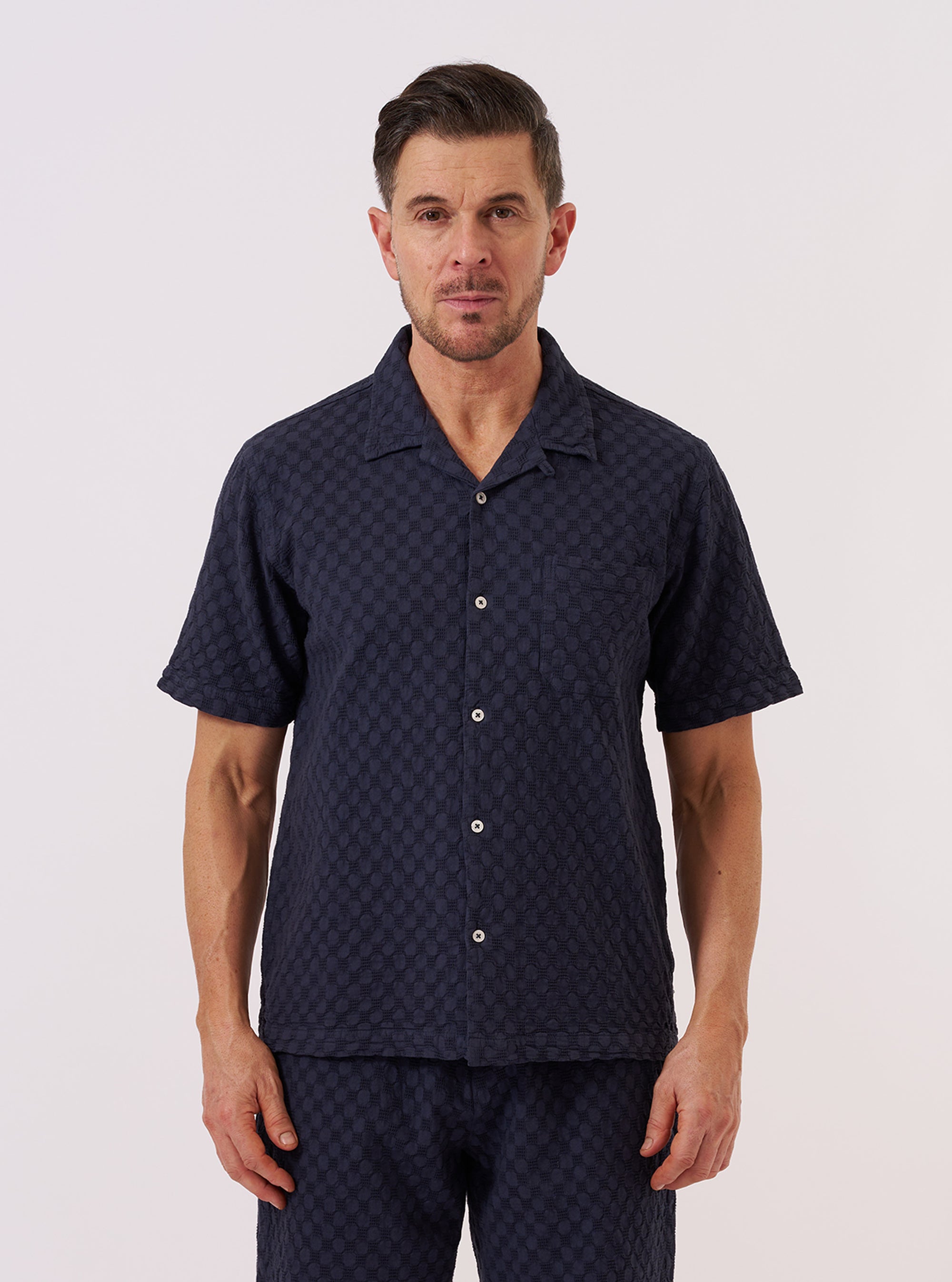A model wears the Navy Dot Waffle Road Shirt against a plain white background. The short-sleeve shirt is shown unbuttoned over a white t-shirt, highlighting its camp collar, single chest patch pocket, and straight hem. The navy blue cotton fabric features a distinct rounded waffle-knit texture throughout.