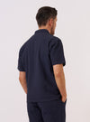 A model wears the Navy Dot Waffle Road Shirt against a plain, neutral background. The shirt features a camp collar, a central button-down front, and a short-sleeve design. The navy blue fabric has a distinct waffle-knit texture with a subtle dot pattern throughout. The image is captured from a rear three-quarter angle, highlighting the shirt's relaxed fit and straight hem.
