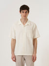 A model wears the Ecru Dot Waffle Road Shirt, styled with dark trousers. The short-sleeve shirt is shown from the front against a plain white background, highlighting its regular fit, single chest patch pocket, and a one-piece camp collar worn open. The ecru cotton fabric features a distinct, textured dot waffle weave.