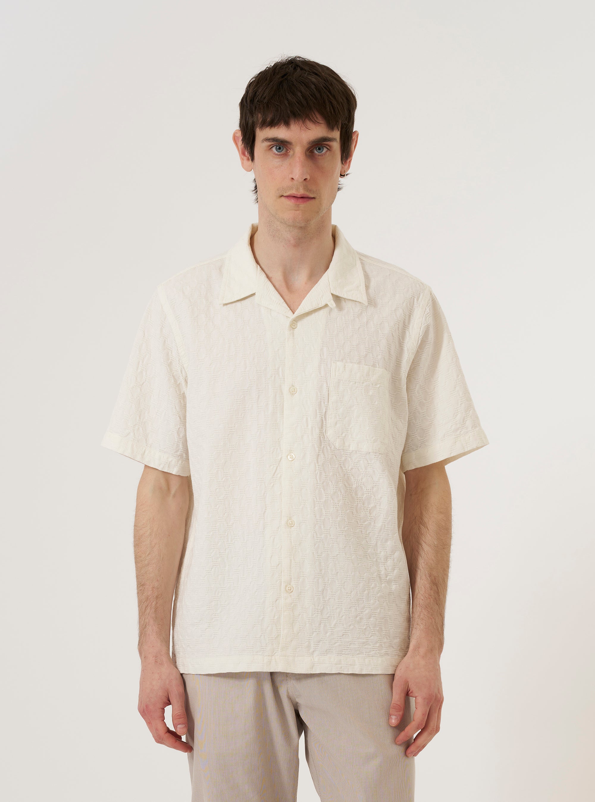 A model wears the Ecru Dot Waffle Road Shirt, styled with dark trousers. The short-sleeve shirt is shown from the front against a plain white background, highlighting its regular fit, single chest patch pocket, and a one-piece camp collar worn open. The ecru cotton fabric features a distinct, textured dot waffle weave.