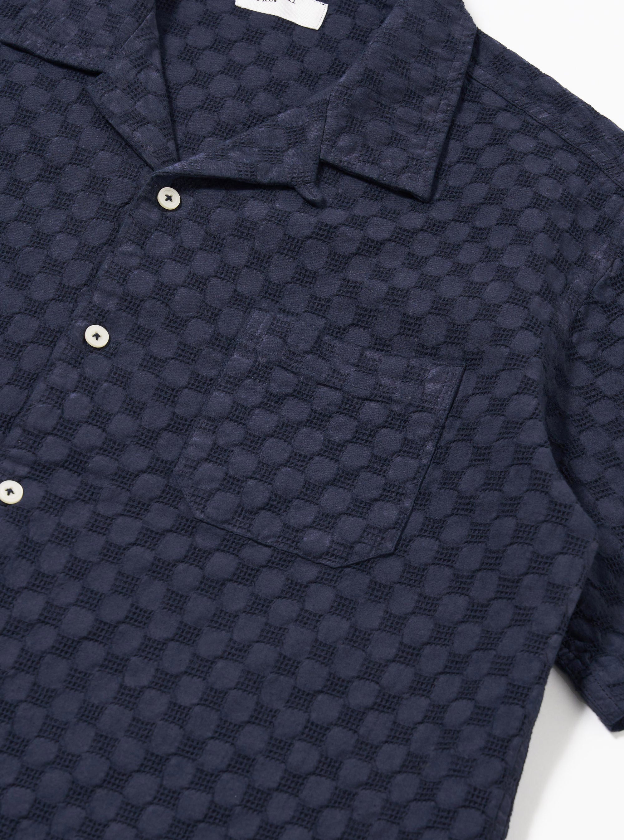 The Navy Dot Waffle Road Shirt is shown from a rear angle as a flat lay against a plain white background. The image highlights the shirt's straight hem, short sleeves, and the subtle textured dot waffle pattern of the navy blue cotton fabric.