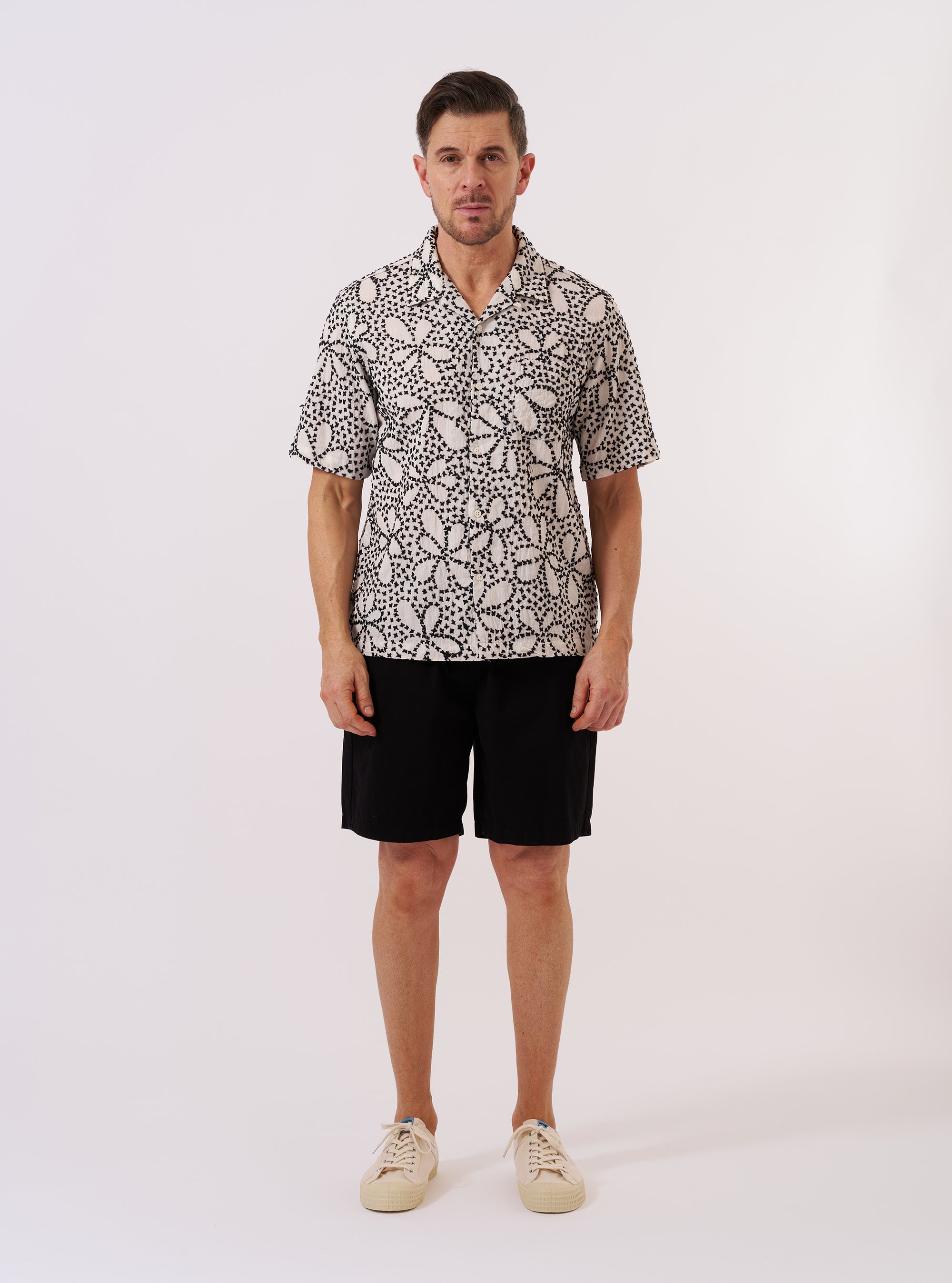 The Ecru/Black Kantha Flower Road Shirt is shown as a flat lay against a plain white background. The shirt features a camp collar, a full-length button front, and a single chest pocket on the left side. The fabric has an all-over intricate black floral pattern on an ecru base, characteristic of the Kantha Flower design.