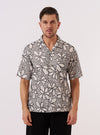 A model wears the Ecru/Black Kantha Flower Road Shirt over a light-colored t-shirt, standing against a plain, neutral background. The shirt features a camp collar, a button-down front, and a prominent black floral print on an ecru-colored cotton fabric. The model is shown from the waist up, highlighting the relaxed fit and short sleeves of the garment.