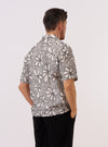 A model wears the Ecru/Black Kantha Flower Road Shirt, shown from the back against a plain white background. The shirt features a relaxed, short-sleeve silhouette with a straight hem and a one-piece collar. The ecru cotton fabric is decorated with an all-over black floral Kantha embroidery, creating a textured, artisanal appearance.
