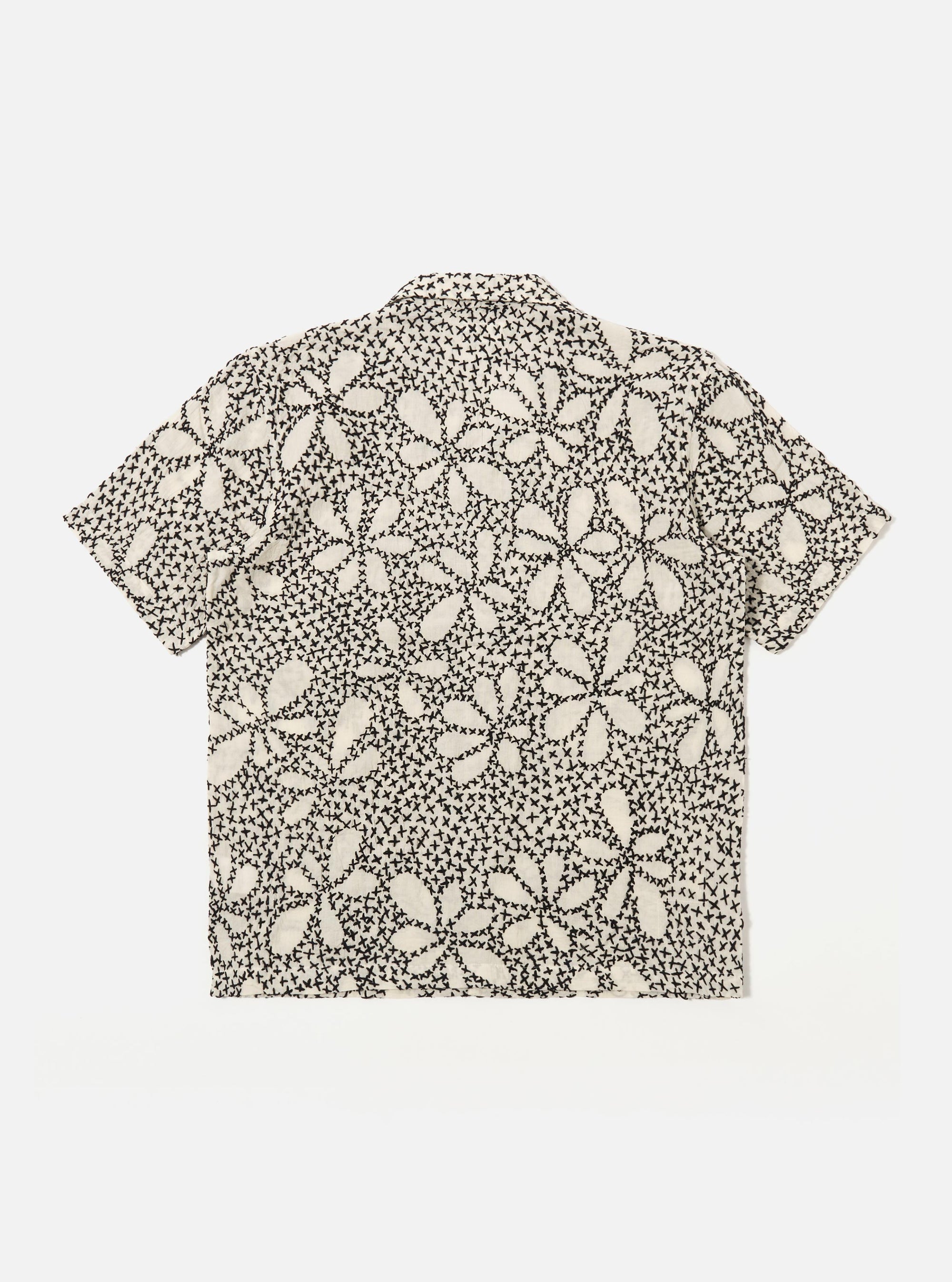The Ecru/Black Kantha Flower Road Shirt is shown from the back as a flat lay against a plain white background. The shirt features an all-over floral pattern created with traditional black Kantha embroidery on an ecru cotton base. The image highlights the straight hem, short sleeves, and the intricate, textured detail of the artisanal hand-stitched embroidery across the entire back panel.