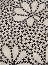 A close-up fabric swatch of the Ecru/Black Kantha Flower Road Shirt highlights the intricate Kantha embroidery on a hand-woven khadi base. The image shows black floral and geometric patterns stitched onto the ecru-colored cotton fabric, emphasizing the artisanal texture and depth of the material.