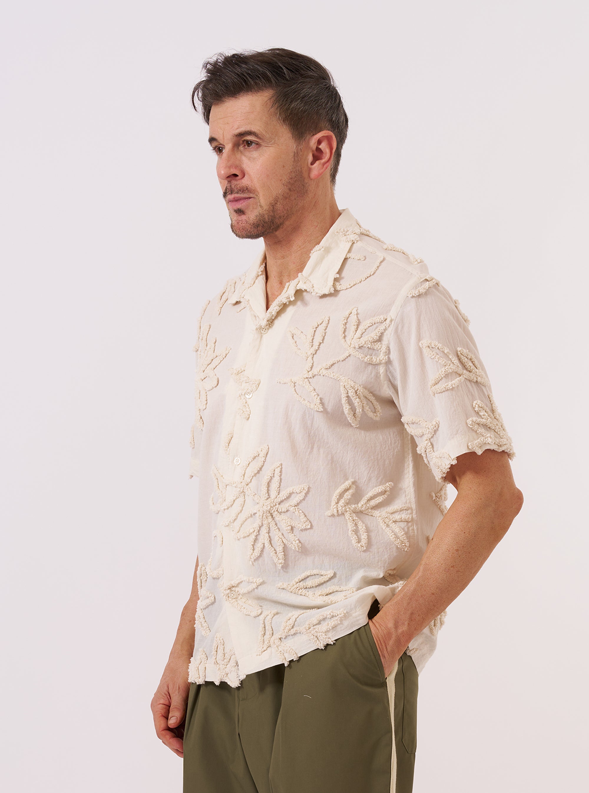 A model wears the Ecru 3D Flower Road Shirt against a neutral background. The short-sleeve shirt features a camp collar with a loop-button closure, a single chest patch pocket, and a straight hem. The ecru cotton fabric is detailed with textured, tone-on-tone 3D floral embroidery throughout.