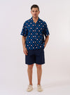 The Indigo Diamond Ikat Deck Shirt is shown as a flat lay against a plain white background. The shirt features short sleeves, a one-piece collar, a five-button front closure, and two large patch pockets at the lower front. The fabric displays an intricate repeating diamond ikat pattern in indigo blue and white.