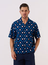 A model wears the Indigo Diamond Ikat Deck Shirt over a light-colored top, standing against a plain white background. The shirt features a distinctive three-color diamond ikat pattern in indigo, a relaxed wide fit, and a clean one-piece collar. Two large patch pockets are prominent on the lower front of the garment, which is shown with the top button undone and the short, loose sleeves visible.